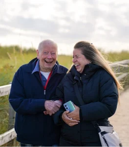 An elderly man and a younger woman walk along a wooden coastal path, laughing together; the woman holds a smartphone.