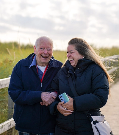 An elderly man and a younger woman walk along a wooden coastal path, laughing together; the woman holds a smartphone.