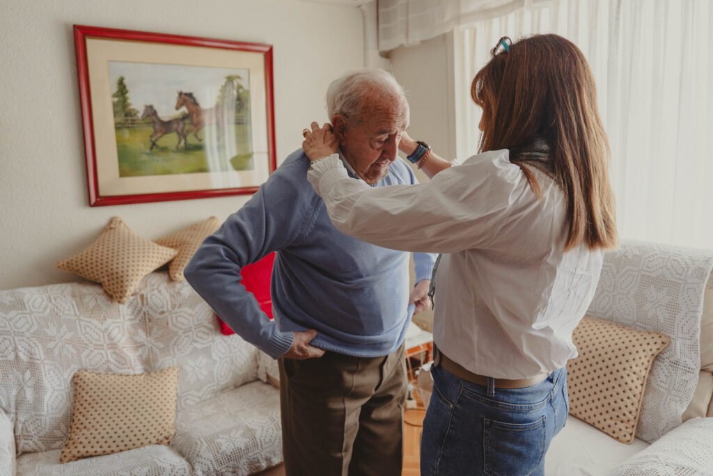 Caregiver helps an elderly man adjust his blue sweater in a cozy living room with a horse painting in the background.