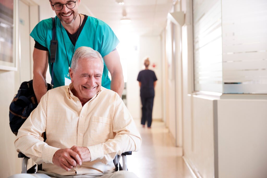 Caregiver in teal scrubs pushes an elderly man in a wheelchair down a bright hospital hallway, both smiling.